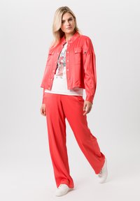 Koraalkleurige, lichte jas met twee voorkruimels, gecombineerd met bijpassende brede broek. Model draagt een grafisch wit t-shirt en witte sneakers.