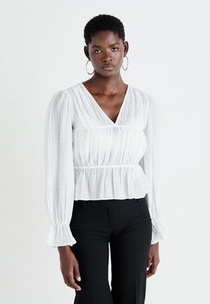 White, textured blouse with a deep V-neck, gathered waist, billowy long sleeves, and flared wrist cuffs. Paired with black pants.