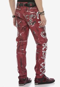 Rote Jeans mit distressed Finish, abstrakten weißen Mustern und Cargo-Taschen. Kombiniert mit schwarzen, mit Nieten besetzten Sneakers. Schwarzer Gürtel mit Logo.