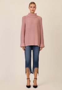 Übergroßer rosa gestrickter Pullover mit hohem Kragen, kombiniert mit verkürzten blauen Jeans mit Fransen am Saum und schwarzen Absatzschuhen.