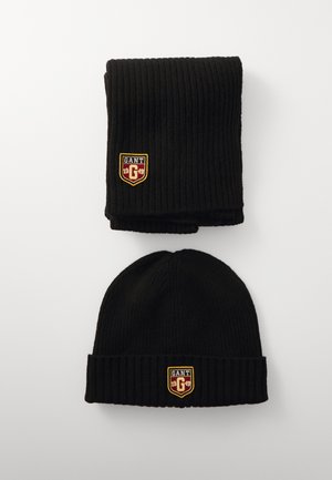 Schwarze gerippte Strickmütze und passender Schal, jeweils mit einem gelben und roten gestickten GANT-Logo-Patch. Weiche, warme Textur.