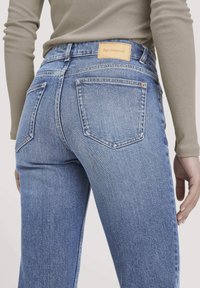 Läheltä kuvattu henkilö, joka pukeutuu vaaleansinisiin farkkuihin, joissa on takataskut, sekä vaaleanbeigeen pitkähihalaiseen paitaan, brändilogo näkyvissä vyötäröllä.