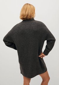 Pull en maille gris foncé surdimensionné avec un col montant, des épaules tombantes et des poignets côtelés. Texture douce et ourlet arrière légèrement allongé.