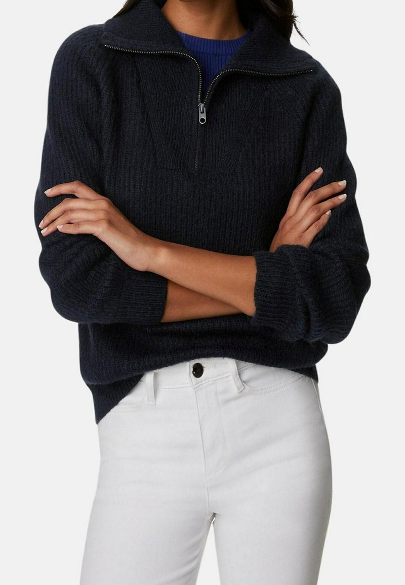 Marineblauw gebreid trui met een kwart rits, geribbeld textuur en een ontspannen pasvorm. Gecombineerd met witte jeans voor contrast.