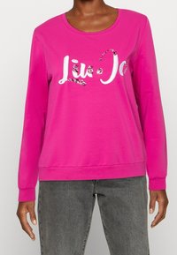 Růžová mikina s dlouhým rukávem, kulatým výstřihem, dekorativním bílým nápisem a ozdobnými prvky pro zvýraznění detailů. Vyrobena z měkké bavlněné směsi.