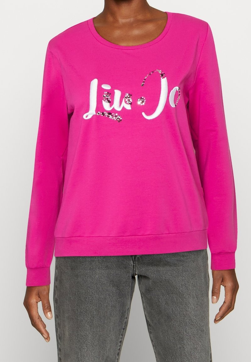 Růžová mikina s dlouhým rukávem, kulatým výstřihem, dekorativním bílým nápisem a ozdobnými prvky pro zvýraznění detailů. Vyrobena z měkké bavlněné směsi.