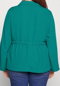 Grön, lättviktig jacka med avslappnad passform, långa ärmar och uppdragen midja. Slät yta och krage utan synlighet framtill.