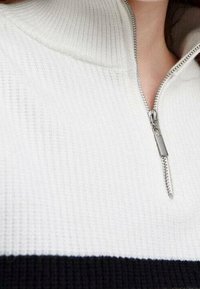 Weißer Reißverschluss-Pullover mit gerippter Textur, einem silbernen Reißverschluss und einem schwarzen horizontalen Streifen nahe dem Saum.