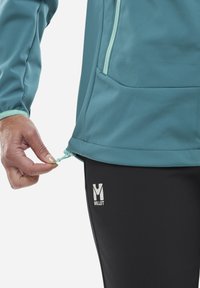 Teeffekt Softshell-Jacke mit Reißverschluss vorne, verstellbarem Saum und Hand in einer schwarzen Hose mit weißem "MILLET"-Logo.