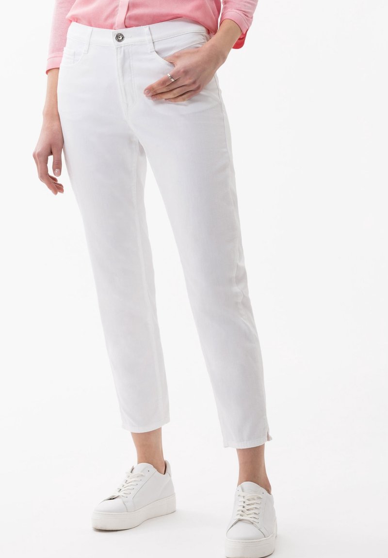 White, slim-fit pants with a smooth texture, front pockets, and a metal button closure. Paired with white, flat-soled sneakers.