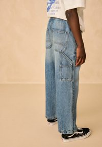 Blugi lejera din denim albastru, cu multiple buzunare, spălare deschisă și textură estompată. Purtați cu adidași negri cu accente albe.