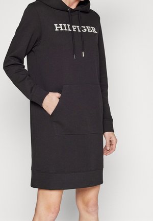 Osoba nosząca czarną sukienkę z kapturem i długim rękawem, z przednią kieszenią i logo "HILFIGER" na piersi.
