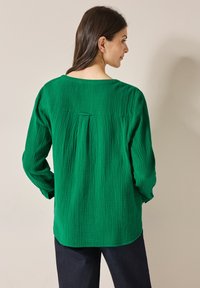Top verde texturizzato con maniche lunghe e scollo a barca. La parte posteriore presenta una plissettatura centrale per un tocco di design in più.
