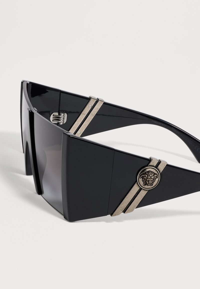 Lunettes de soleil à visière noire oversize avec de larges branches, comportant des doubles bandes argentées et un médaillon rond avec une tête de Méduse sur les côtés.