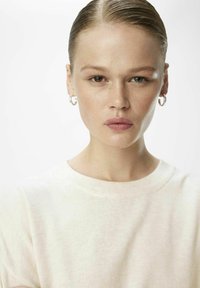 Lys beige strikket sweater med rund halsudskæring. Modellen har kort, slicket tilbage hår og bærer små sølv ringeøreringe.
