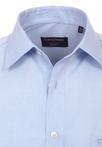 Lichtblauw overhemd met een moderne pasvorm, voorzien van een puntige kraag, witte knopen en een klein borstzakje. Etiket leest 'CASA MODA PREMIUM EDITION.'