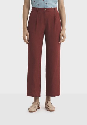 Kobieta ubrana w wysokie spodnie o szerokich nogawkach w kolorze rdzy, zapinane na guzik i suwak z przodu, w połączeniu z beżowymi plecionymi sandałami.