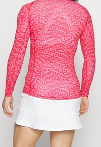 Bakifrån syns en person som bär en åtsittande långärmad röd topp med ett vitt geometriskt labyrintmönster och en vit veckad kjol.