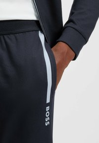 Marineblauwe sport joggers met een lichtblauwe zijstreep en een logo-opdruk. De stof heeft een gladde textuur en een elastische tailleband.