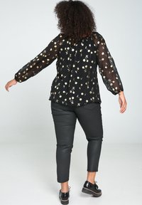 Blouse noire transparente avec motif floral doré, manches ballon, encolure élastique ; associée à un pantalon noir ajusté et des chaussures noires à lacets.