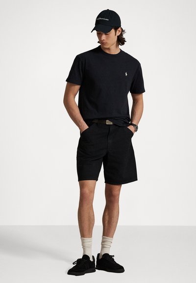 Svart bomull T-skjorte med logo, kombinert med svarte shorts. Antrekket inkluderer svarte sneakers og kremfargede sokker, samt en svart caps.