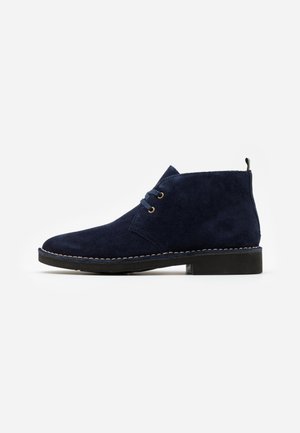 Bottines en daim bleu marine avec un bout arrondi, des accents de couture et deux œillets pour les lacets. Semelle en caoutchouc avec un léger talon et une boucle de traction.