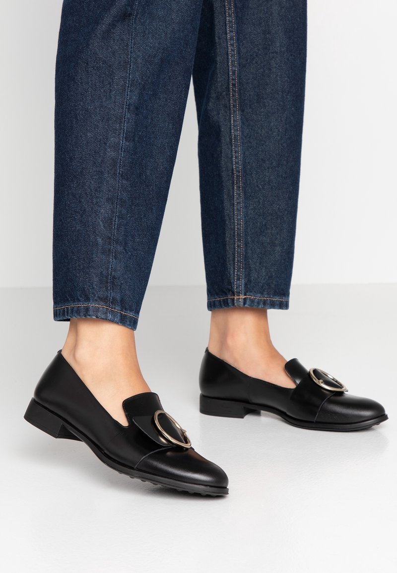 Pieds en mocassins en cuir noir avec de grandes boucles argentées, associés à un jean denim court bleu foncé sur un fond clair uni.