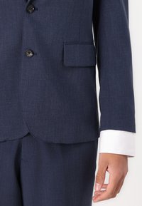 Donkerblauw colbert met een borstzak, twee zwarte knopen en witte manchet details. De stof heeft een subtiele textuur en een getailleerde pasvorm.