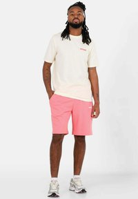 Lichtbeige T-shirt met rood logo, gecombineerd met roze shorts. De outfit heeft een ontspannen pasvorm, zachte stof en een eenvoudig ontwerp.