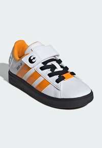Baskets blanches avec des rayures orange, lacets noirs et une sangle Velcro. Comprend le logo BB-8, une semelle en caoutchouc noir et une tige texturée.