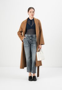 Femme portant un long manteau marron, un haut translucide bleu marine, un jean gris taille haute, des mocassins noirs et tenant un petit sac à main blanc.