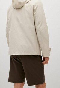 Giacca cappuccio beige chiaro con maniche lunghe, tessuto testurizzato e polsini abbottonati, abbinata a pantaloni corti marrone scuro. Vista posteriore mostrata.