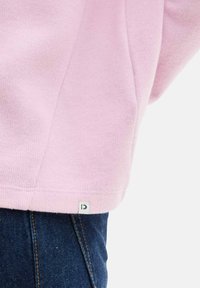 Svetloružový skrátený sveter s rebrovanou textúrou a malou látkovou nášivkou na dolnom leme, v kombinácii s tmavomodrými džínsami.