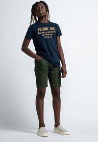 Granatowa koszulka z żółtym napisem, zielone spodenki i jasne buty. Koszulka ma luźny, swobodny krój i wygodny materiał.