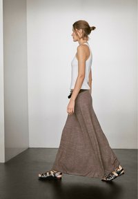 Witte tanktop en een lange, bruine, gestructureerde rok, gecombineerd met zwarte sandalen met meerdere banden, lopend tegen een neutrale achtergrond.