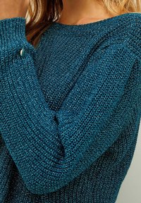 Close-up of a person wearing a shiny, textured teal knit sweater with ribbed cuffs and a small silver button detail.