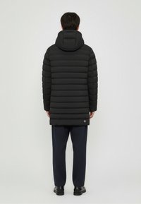 Black puffer jacket with horizontal quilted sections, hood, and branded patch at the hem. Worn over dark trousers and black shoes.