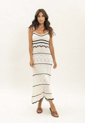 Femme aux longs cheveux bruns ondulés portant une robe maxi sans manches en maille à motifs blancs et noirs et des sandales marron, debout et regardant devant elle.