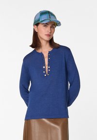 Blauwe langarmshirt met een diepe V-hals, voorzien van gouden knopen aan de voorkant. Gladde stof, gecombineerd met een glanzende bruine rok.