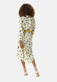 Floral midi dress in white, featuring yellow and black patterns, long sleeves, and a cinched waist with a yellow belt. Paired with white strappy heels.