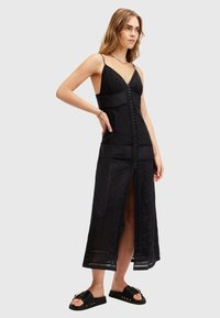 Black maxi dress with adjustable spaghetti straps, intricate embroidered pattern, button-up front, and side slit. Paired with black sandals.