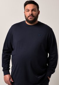 Man med skägg som bär en enkel mörk marinblå långärmad skjorta, står med en hand i fickan mot en ljus neutral bakgrund.