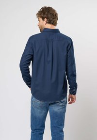 Jūrų mėlynos mygtukų marškiniai su klasikiniu apykakle, ilgomis rankovėmis ir išlenktu apačiu, derinami su šviesiai mėlynomis džinsinėmis kelnėmis, turinčiomis užpakalines kišenes.