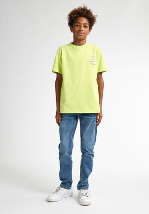 Neon groene katoenen T-shirt met korte mouwen en ronde hals, voorzien van een grafisch ontwerp in het blauw nabij de borst, gecombineerd met blauwe spijkerbroek en witte sneakers.