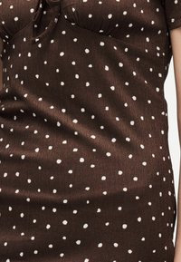 Καφέ φόρεμα από ύφασμα με μικρές λευκές πουά και υφή με μοτίβο, με μικρό άνοιγμα και κορδόνι δεμένο κοντά στη γραμμή του λαιμού.
