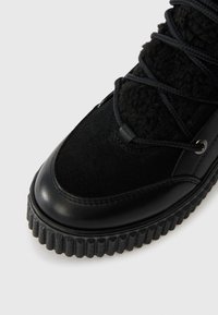 Černá zimní obuv s tlustou rýhovanou podešví, svrškem z nubuku a kůže, s jazykem z ovčího rouna a překříženým šněrováním na bílém pozadí.