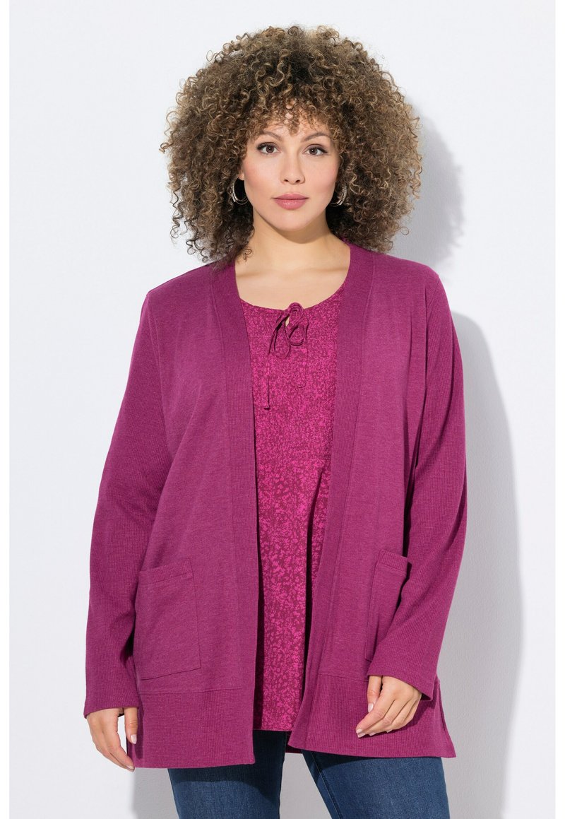 Magenta offene Strickjacke mit langen Ärmeln, zwei vorderen Taschen und strukturiertem Strick. Über einem passendem gemusterten Oberteil getragen.