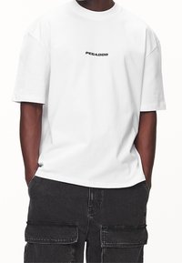 Bílý bavlněný tričko s krátkým rukávem a kulatým výstřihem, s černým textovým logem "PEGADOR" umístěným uprostřed na hrudi.