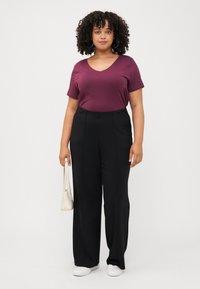 A person with curly hair stands confidently, wearing a burgundy top, black wide-leg pants, and holding a small white bag.