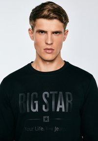 Junger Mann mit kurzen braunen Haaren, der einen schwarzen Sweatshirt trägt, auf dem vorne "BIG STAR Your Life. Your Jeans." gedruckt ist, neutraler Hintergrund.
